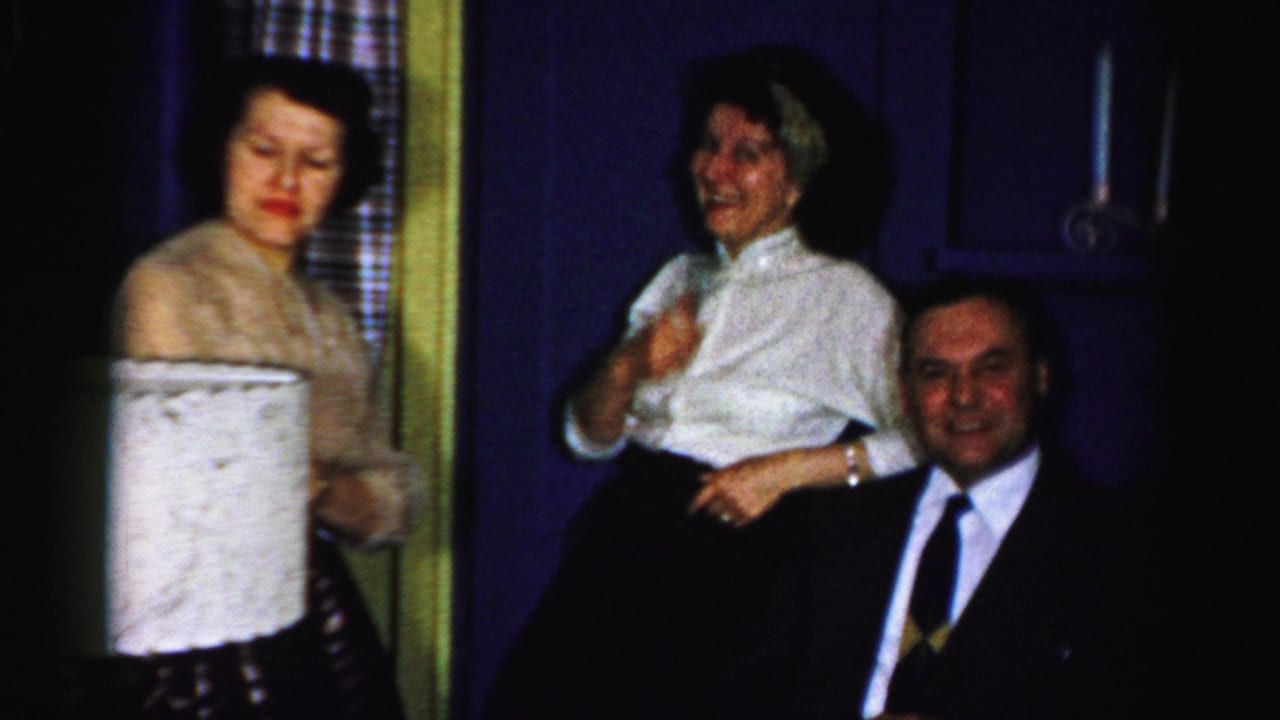 Albany 1956 party frame with two women standing near a seated man in a suit, with a large lampshade in the foreground.