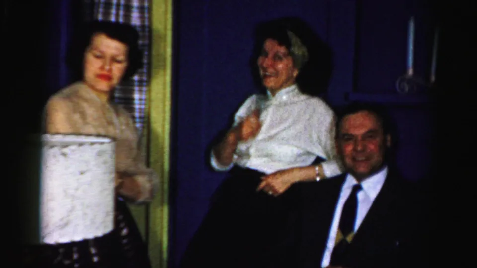 Albany 1956 party frame with two women standing near a seated man in a suit, with a large lampshade in the foreground.