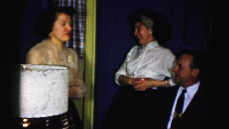Albany 1956 home-party frame showing adults gathered near a lamp, with saturated blue walls and soft 8mm grain.