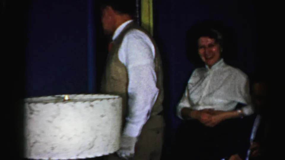 Albany 1956 indoor party frame with a standing man seen from behind and a smiling woman near the wall and lamp.