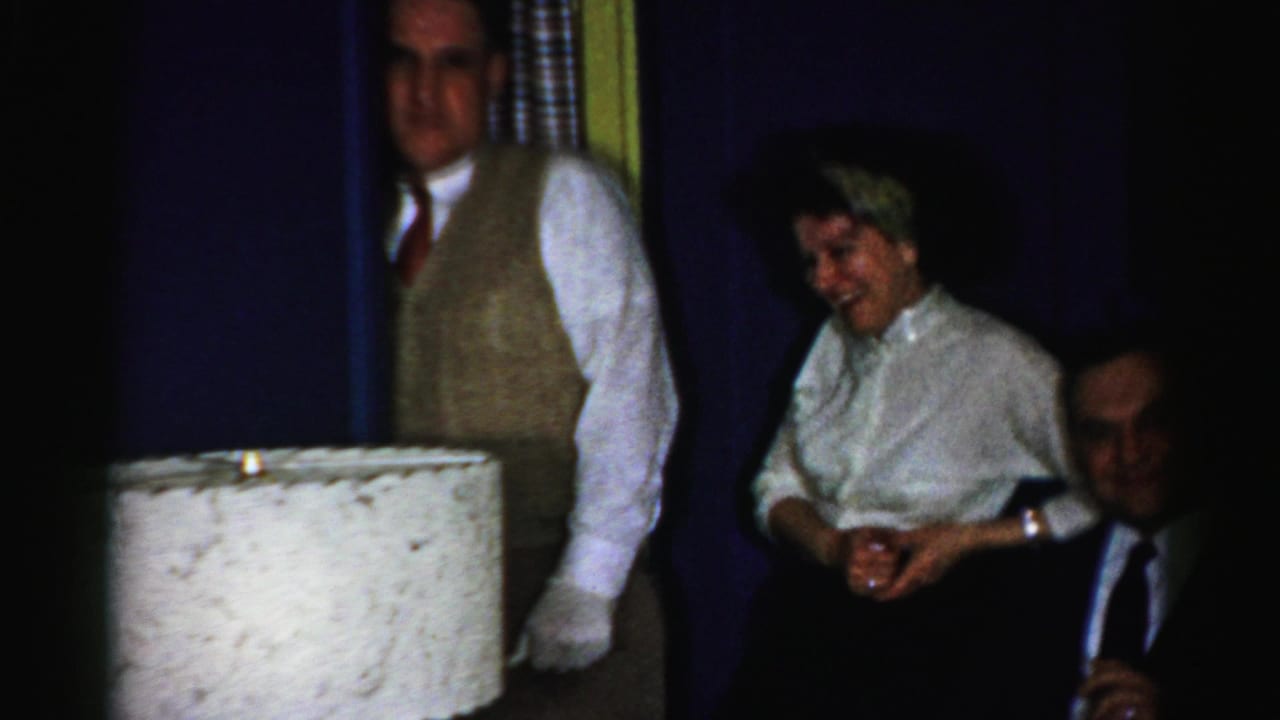 Man in vest and white gloves stands beside a laughing seated woman and a man in a dark suit.
