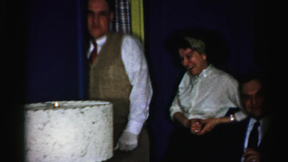 Albany, New York home-movie frame from 1956 showing a gloved man in a vest with seated adults laughing in a dark interior.