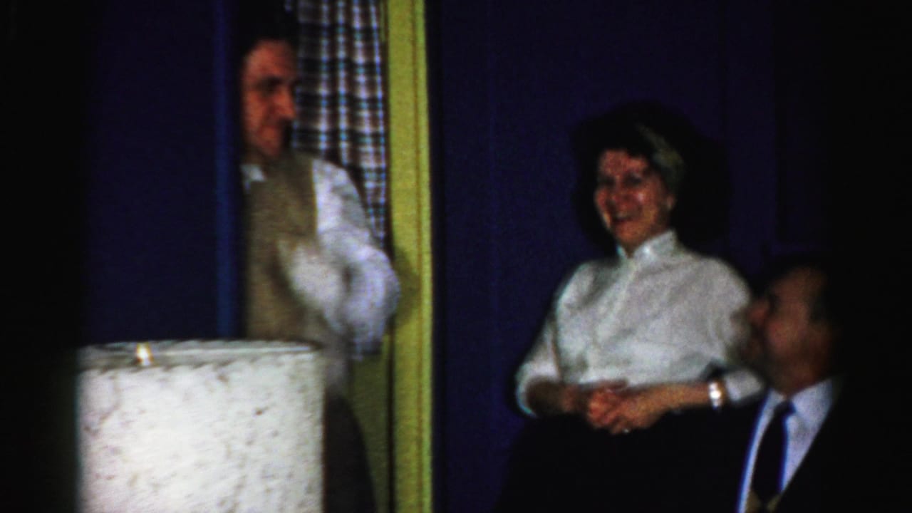 Gloved man gestures near a smiling woman and seated man in a dark suit.