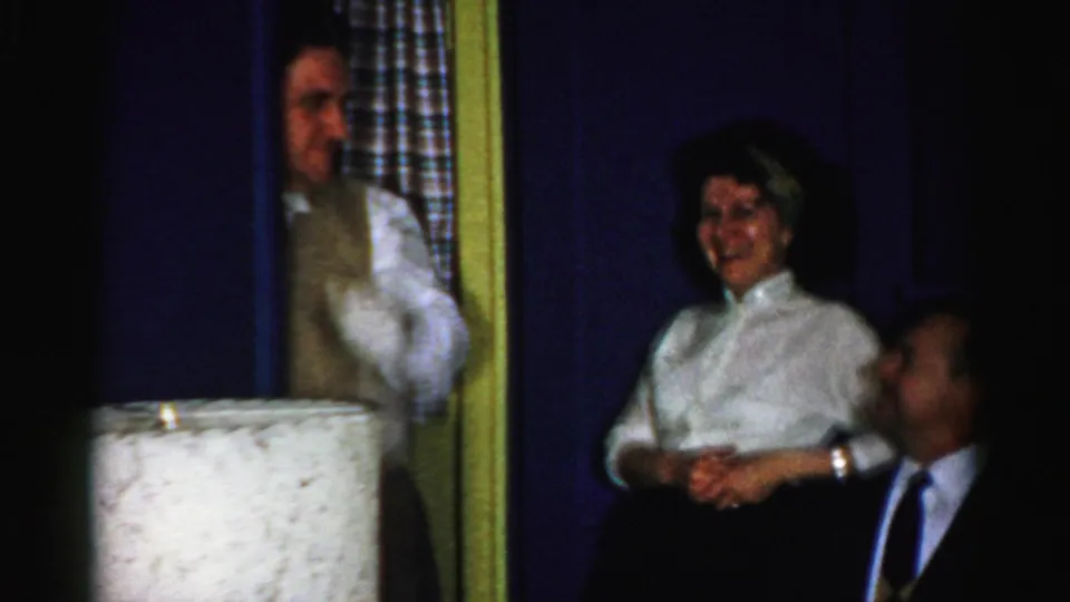 Albany, New York 1956 party frame with a man in white gloves gesturing as a woman smiles beside a seated man.