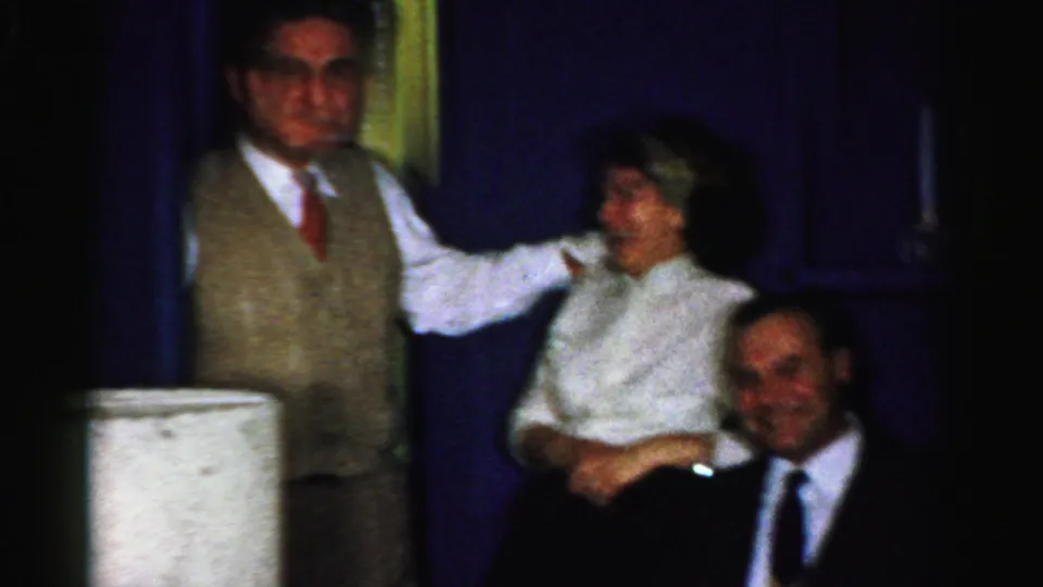 1956 Albany home-movie frame showing a vested man facing the camera as a woman laughs in a private indoor gathering.