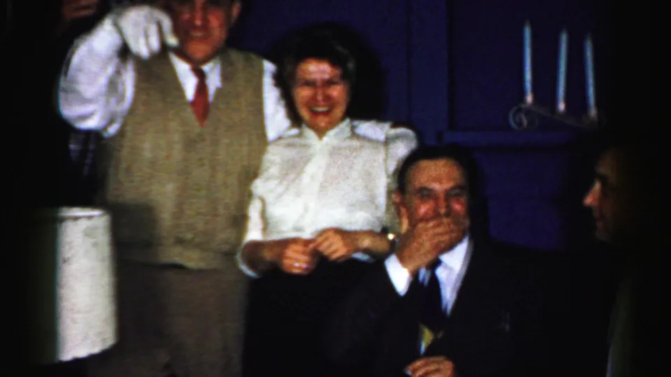 Albany, New York 1956 frame of adults laughing indoors, with a gloved man standing behind a woman and a seated man covering his mouth.