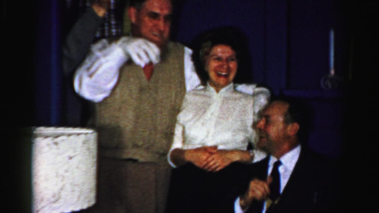 1950s Albany home-movie frame of adults laughing together indoors, with a man in a vest, a woman in a white blouse, and a seated man in a dark suit.