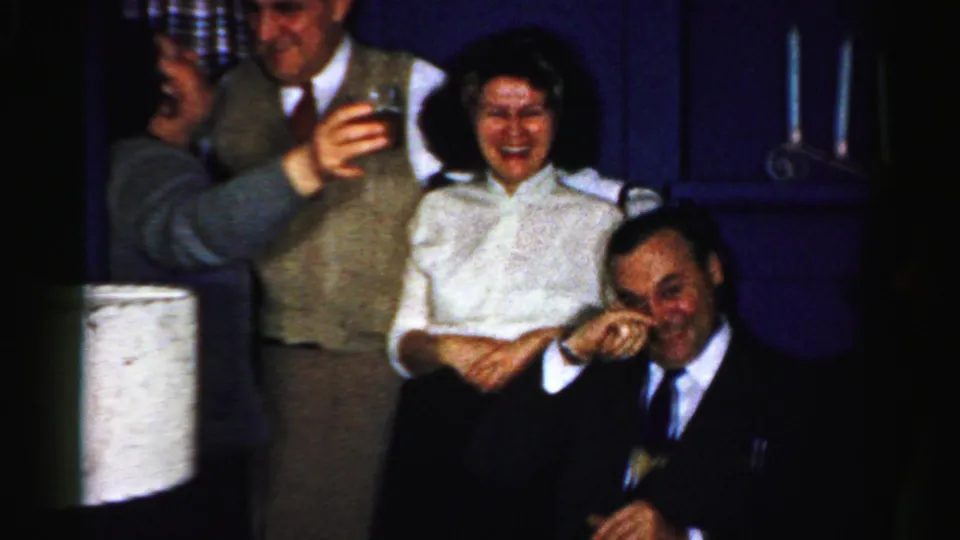 A candid 1950s Albany home-movie frame shows laughing adults indoors while a seated man in a suit wipes or covers his face during the gathering.
