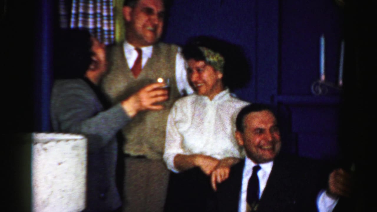 A 1956 Albany home-movie frame shows adults laughing and turning during a toast, with a seated man in a dark suit smiling in the foreground.