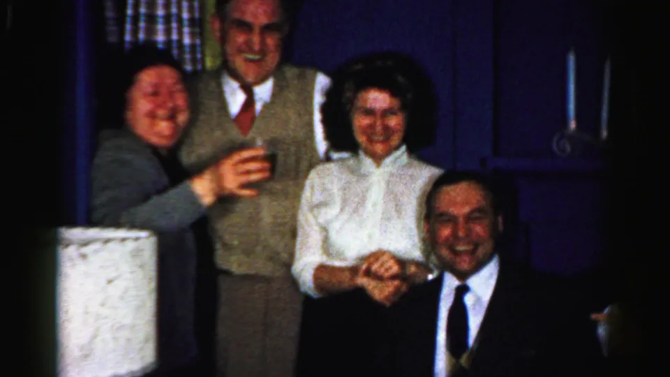 This 1956 Albany party frame shows three standing adults and a seated man smiling, with raised glasses and a blue interior backdrop behind them.