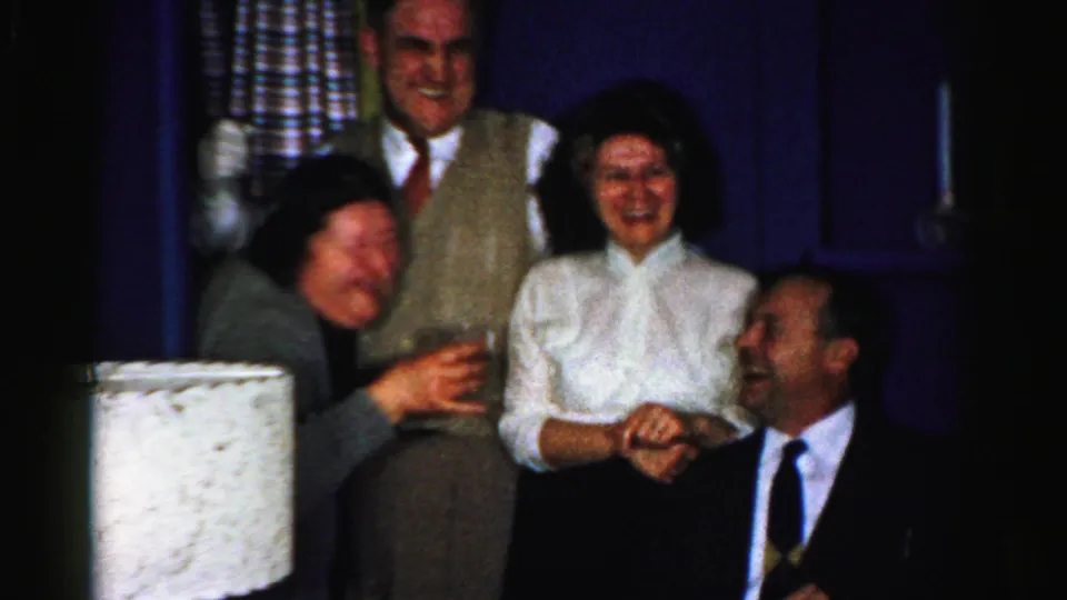 A 1956 Albany home-movie frame captures adults laughing at an indoor party, with a woman holding a drink and a seated man smiling up from the foreground.