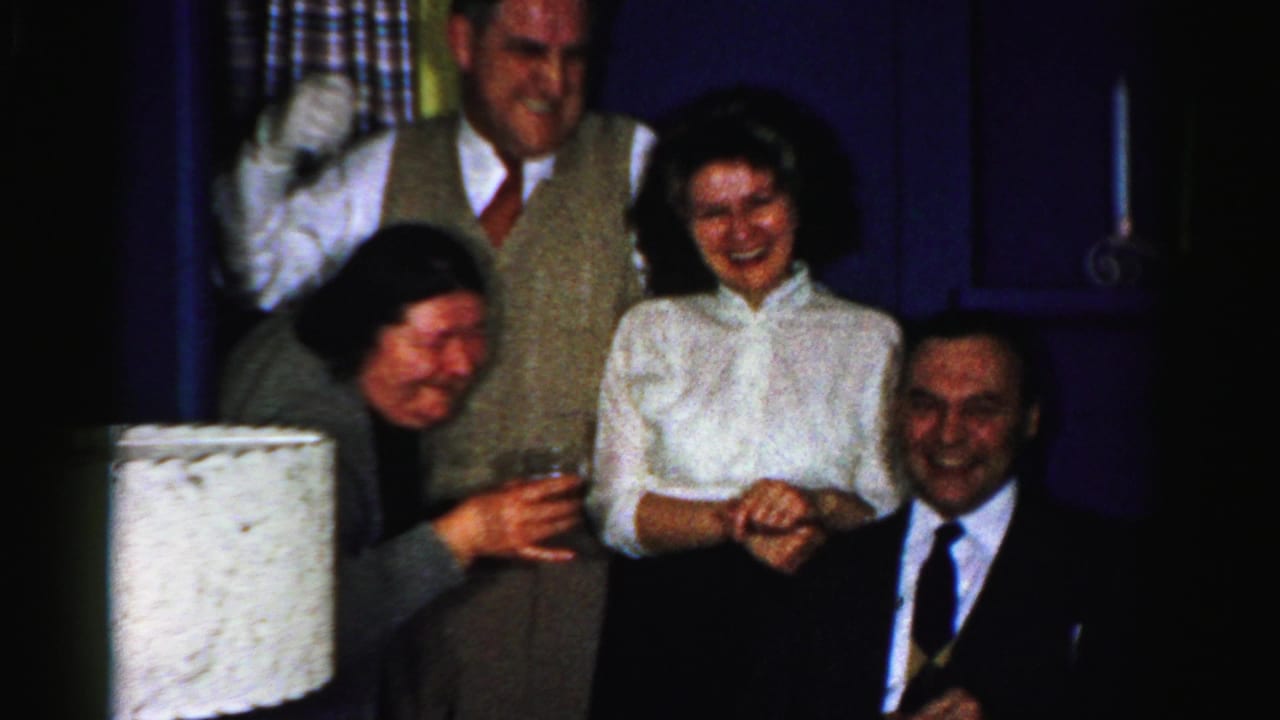 Adults gathered indoors, smiling and holding drinks near a table lamp.