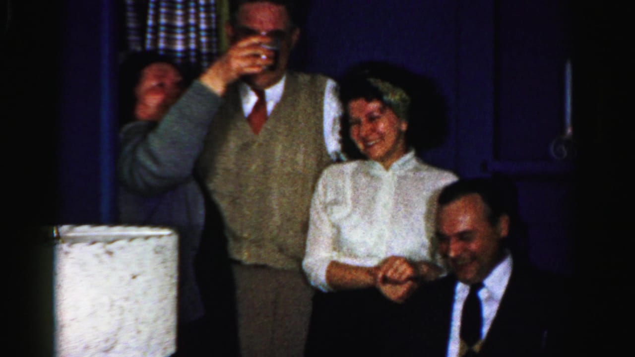 Adults in a dim room laugh as one person drinks from a glass.