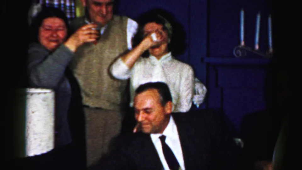 1950s Albany home-movie frame with adults raising glasses around a seated man in a suit during a family party.