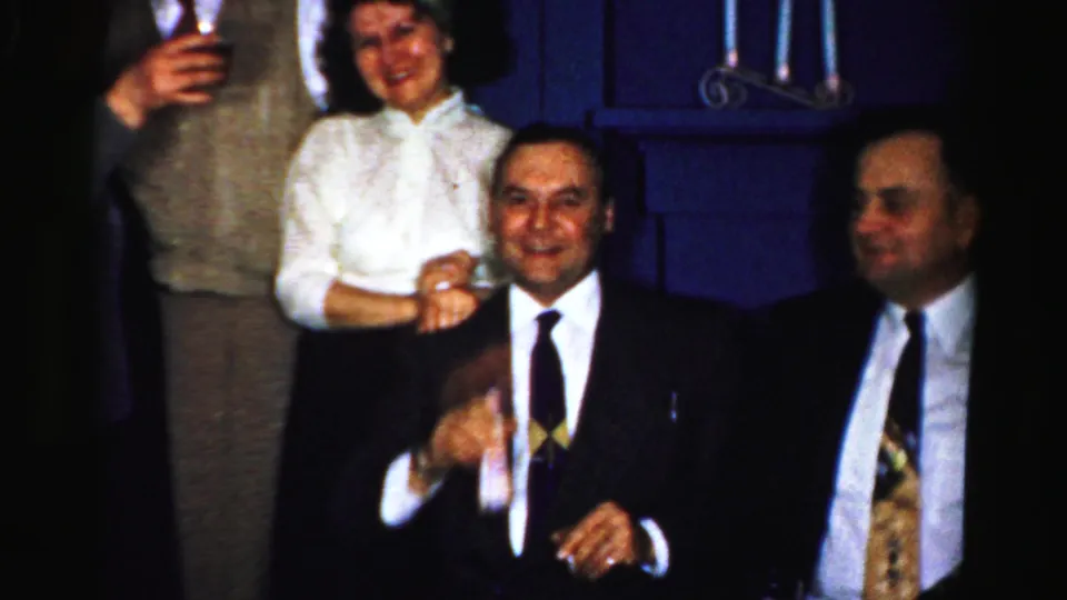 1950s Albany home-movie frame showing two seated men and standing adults smiling with drinks at an indoor gathering.
