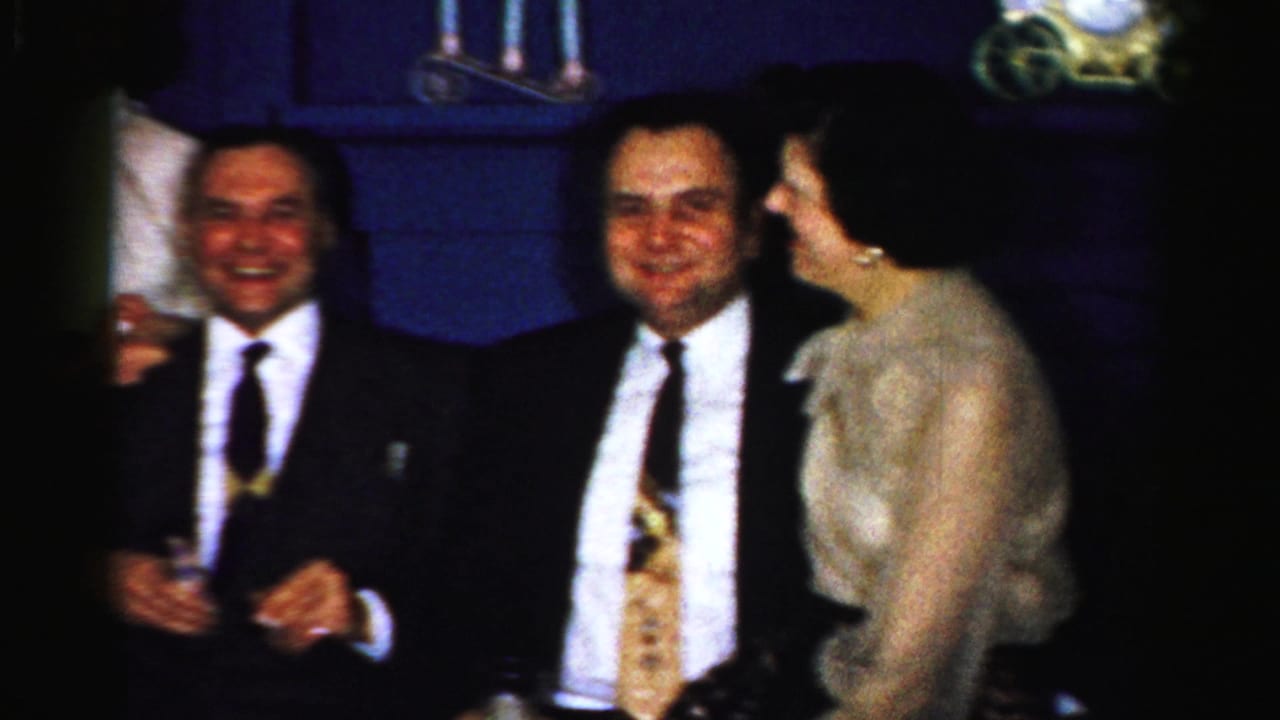 Two men in dark suits sit with a woman in a light dress in a dim room.