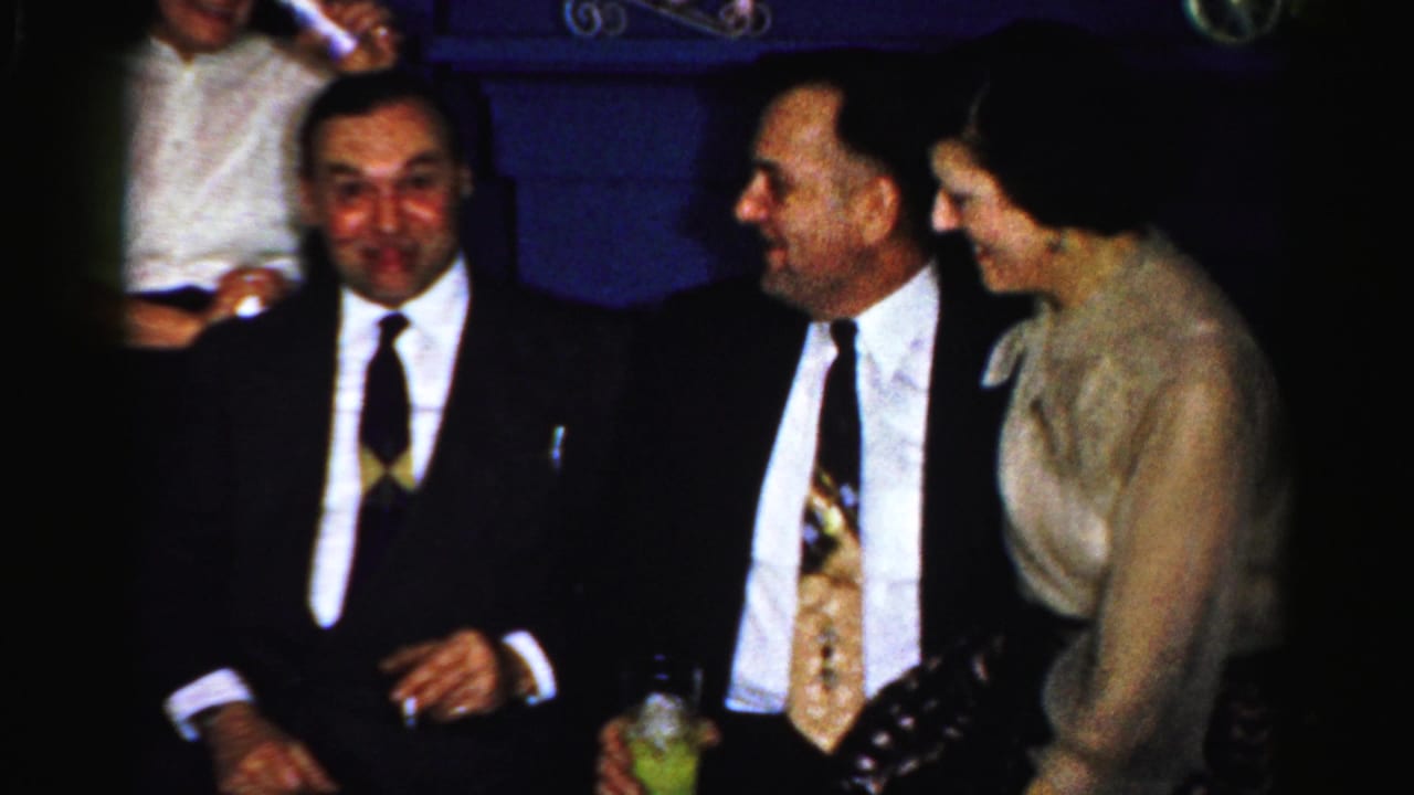 Two men in suits and a woman in a light dress sit together with drinks nearby.