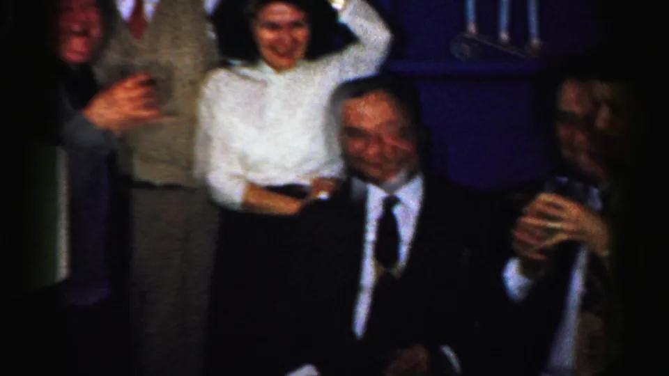 Adults in formal clothing cluster in a dark Albany room, including a woman holding a drink and a man in a white shirt.