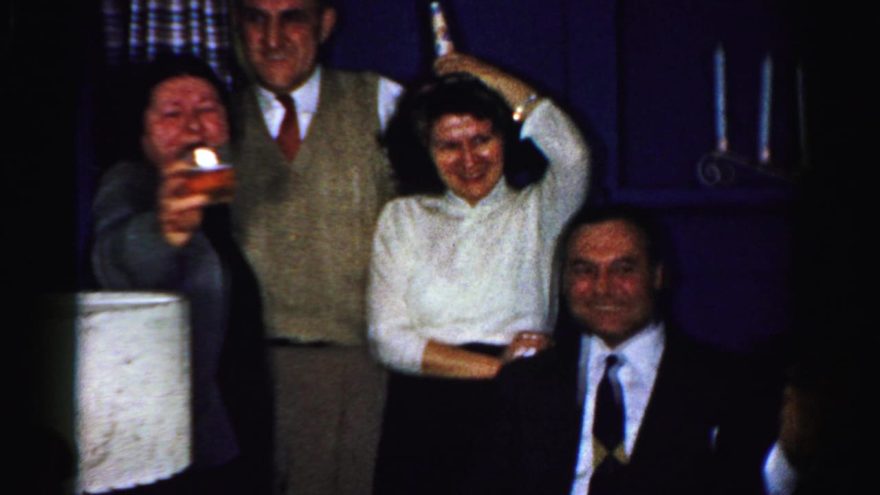 Albany home-movie frame from the 1950s showing adults in formal clothes posing in a dim living room as one woman holds up a drink.