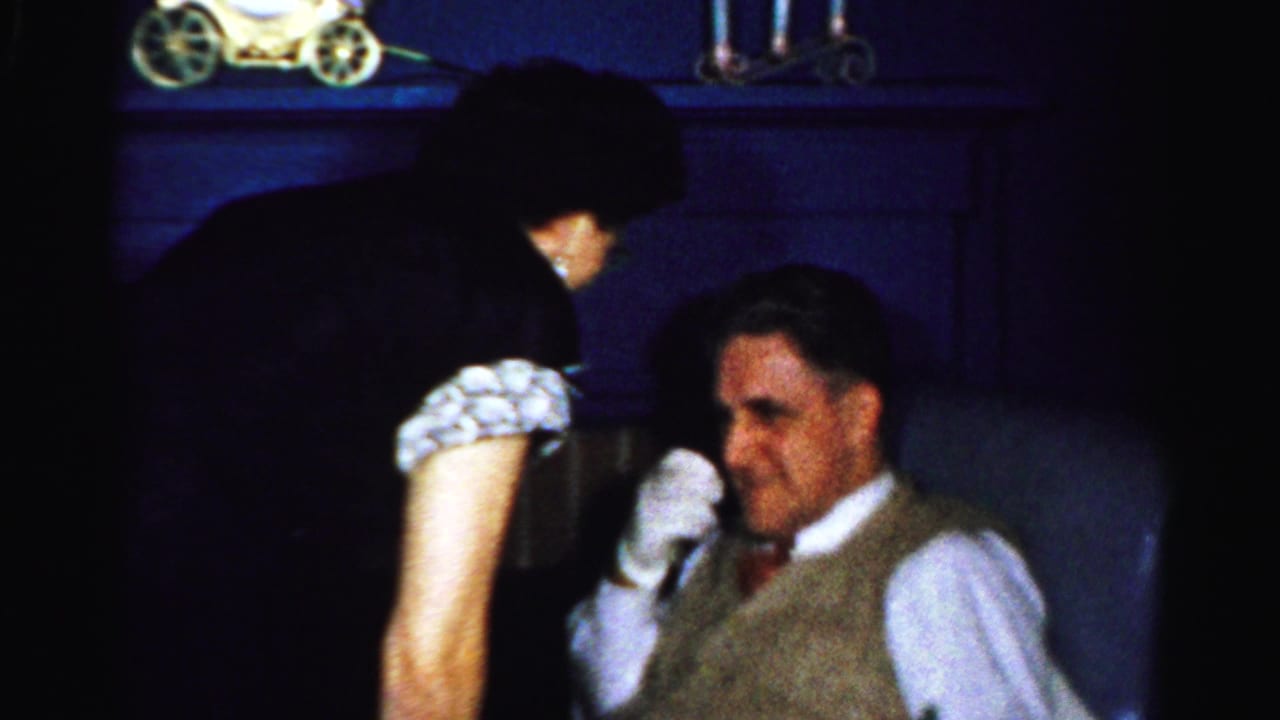 1950s Albany home-movie frame showing a woman leaning close to a seated man in a vest and tie beside a dark mantel.