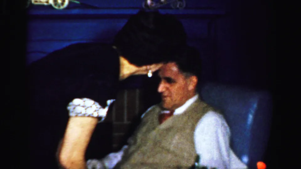 Albany 1950s home-movie frame with a woman leaning beside a seated man in dress clothes during an indoor party.