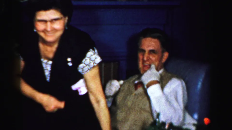 1950s Albany party frame showing a smiling woman standing by a seated man in a vest inside a private living room.