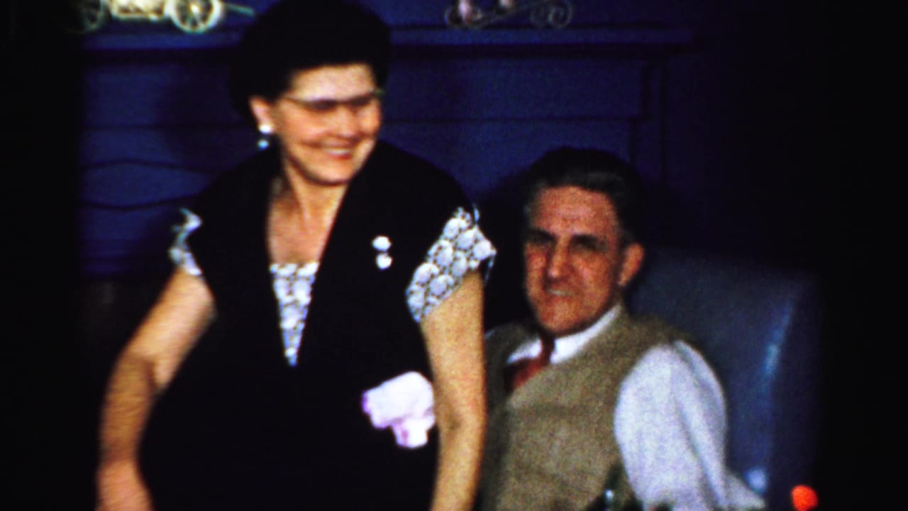 In Albany 1956 home-movie footage, a woman stands beside a seated man in a blue chair as both appear amused.
