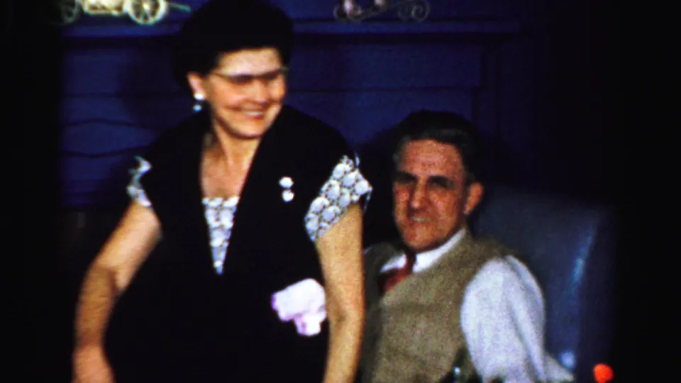 In Albany 1956 home-movie footage, a woman stands beside a seated man in a blue chair as both appear amused.