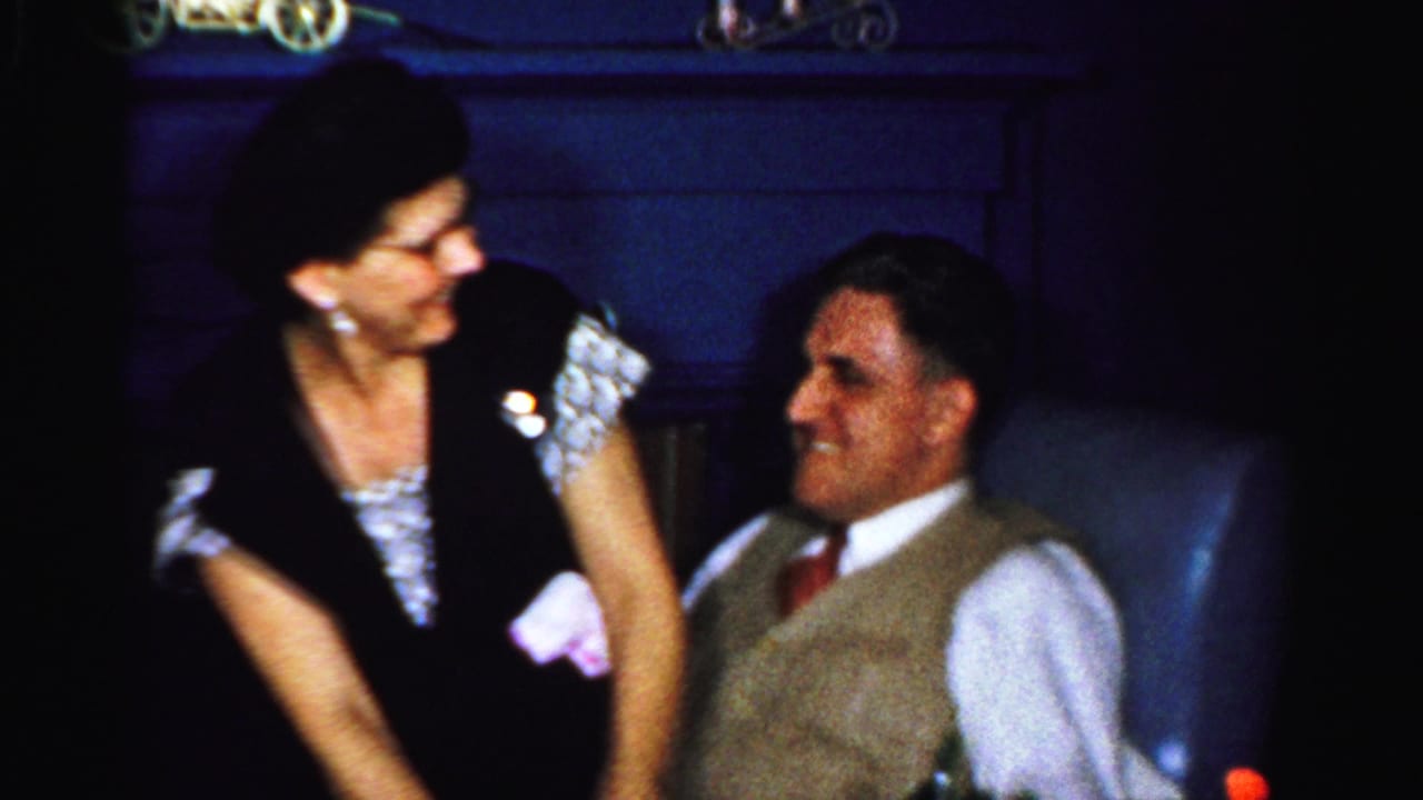 The woman looks toward the seated man in a blue chair, with midcentury clothing visible in the dim Albany interior.