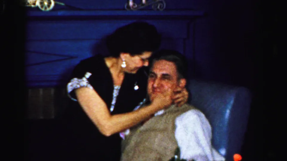 In the 1956 Albany home movie, the woman leans in and touches the seated man's face during an affectionate exchange.