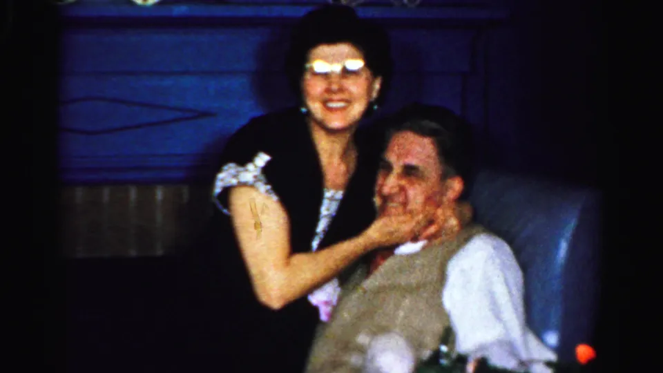 The woman smiles broadly while holding the seated man's face, with the blue chair and dark interior visible behind them.