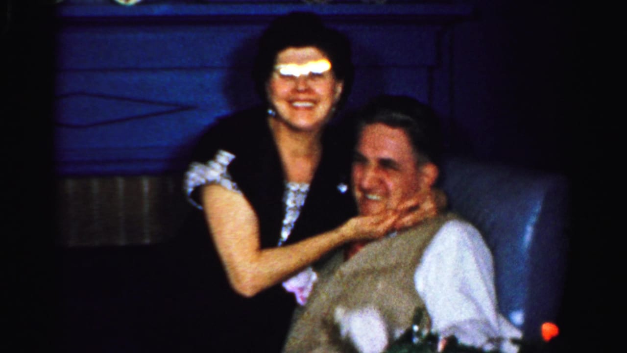Woman in glasses smiles while holding a seated man's face on a dim couch.
