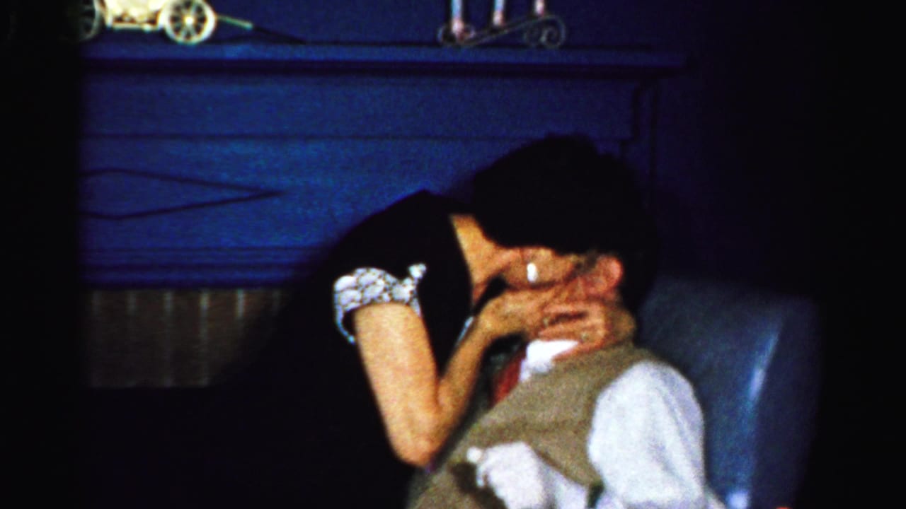 The woman leans toward the seated man for a kiss, with the dark interior and couch visible around them.