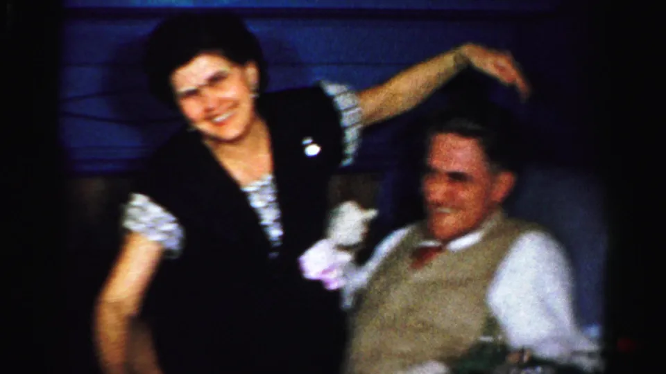 The woman stands and gestures beside the seated man, who smiles faintly in the dim party room.