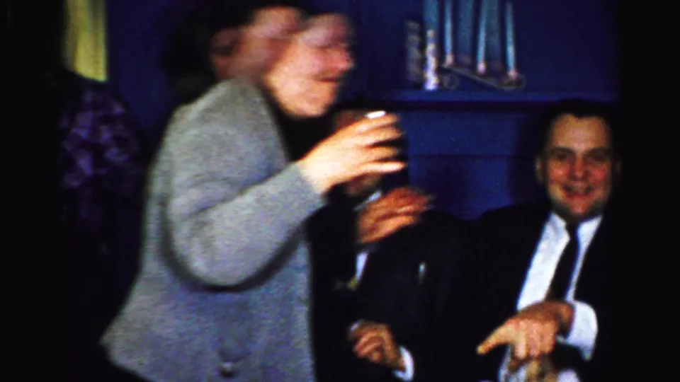Two standing adults with drinks pass in front of a seated man in a suit during the Albany home gathering.