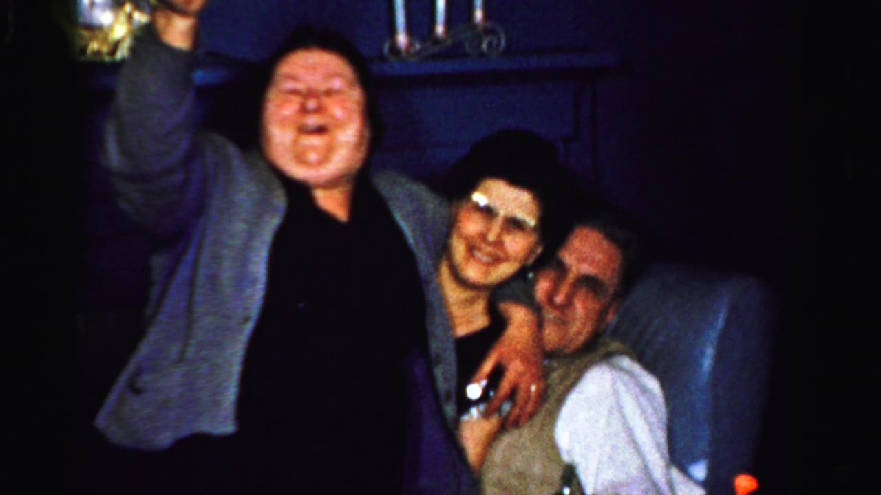 In a 1956 Albany home-movie frame, three adults gather closely in a dark room while a woman in a gray cardigan raises a glass.