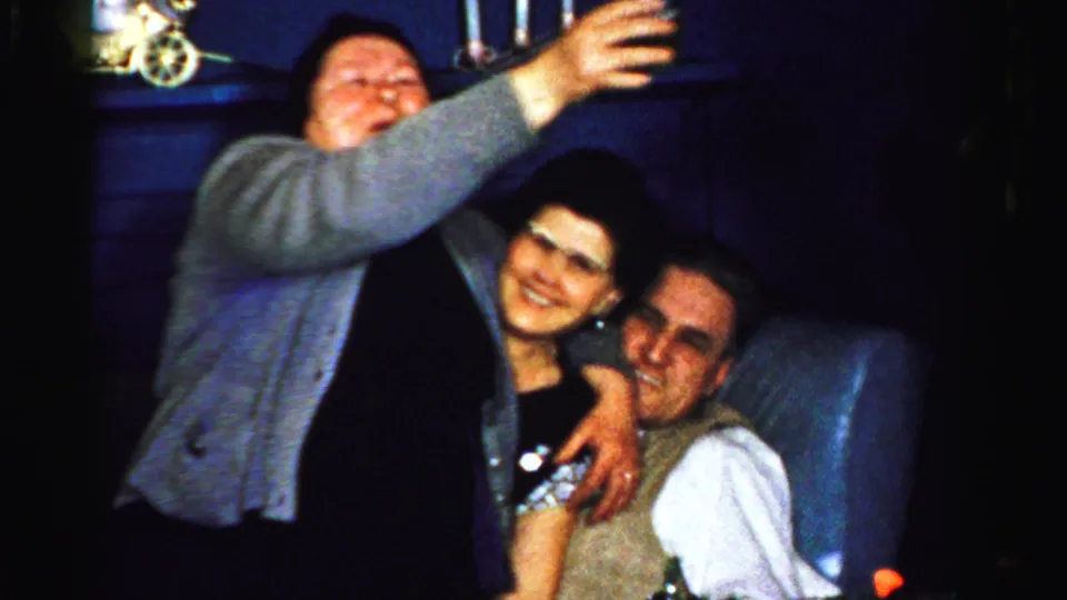 In the Albany home-movie party scene, a woman stretches an arm forward with a drink as two adults sit close together behind her.