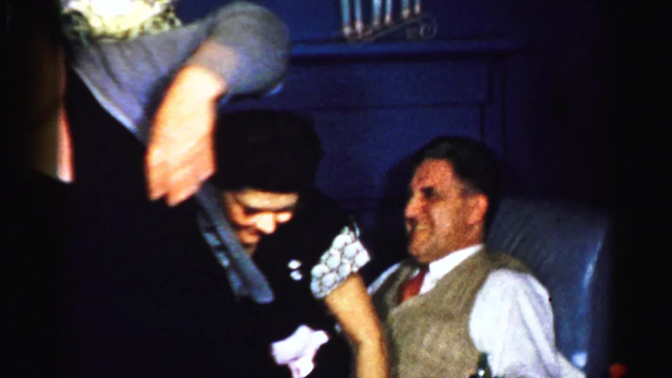 A dim 1956 Albany interior frame shows adults gathered around a seated man in vest and tie as the celebratory motion shifts.