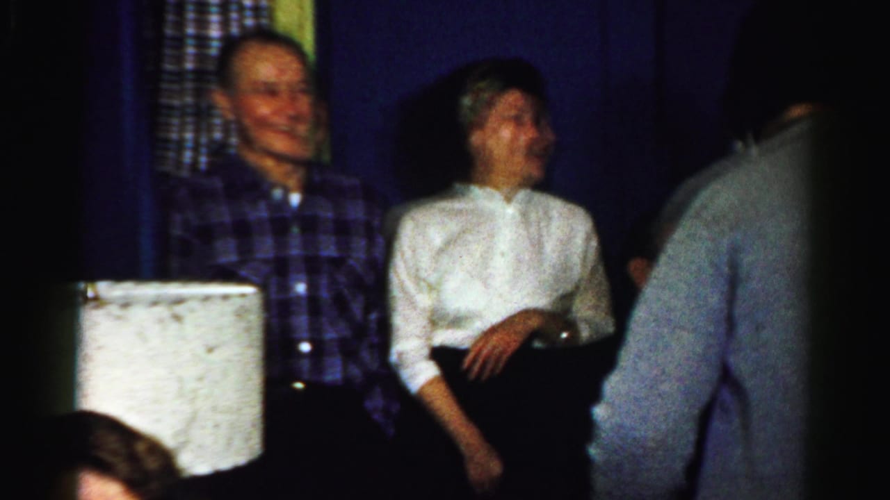 Adults laughing and talking in a dim room with a lamp shade in the foreground.