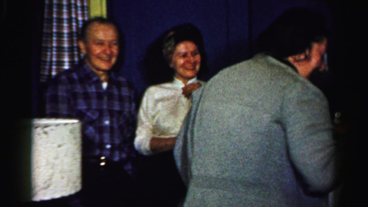 1950s Albany home-movie frame with smiling adults near patterned curtains as a guest in a gray sweater moves through the foreground.