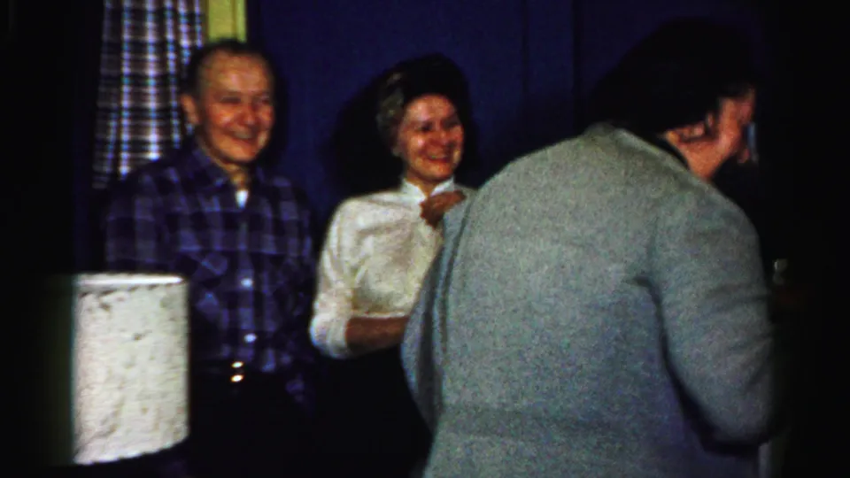 1950s Albany home-movie frame with smiling adults near patterned curtains as a guest in a gray sweater moves through the foreground.