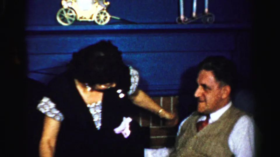 1950s Albany home-movie frame of a woman leaning toward a seated man in a domestic interior with dark paneling and small decorative objects.