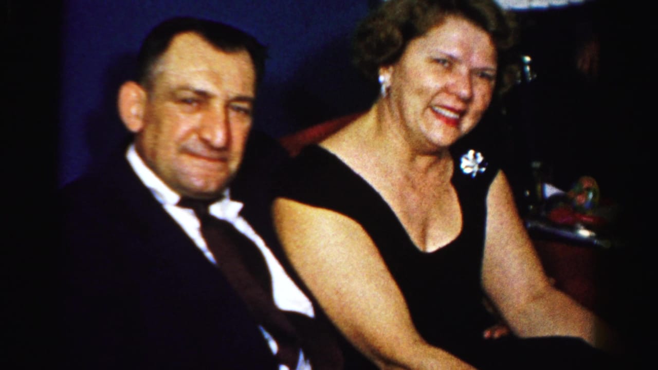 A formally dressed couple sits close together in a dim Albany interior, smiling toward the 8mm camera.