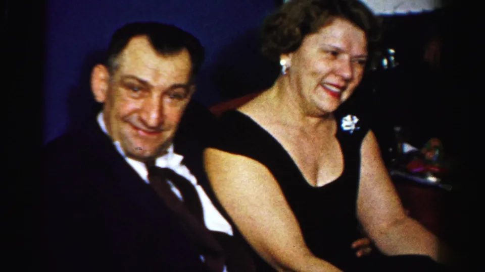 The seated couple remains in view, with the woman’s brooch and black dress clear in the 1950s home-movie frame.