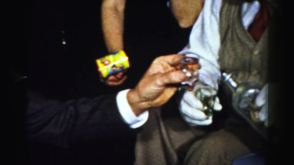 Close party detail shows an adult hand raising a small glass while another guest holds a yellow film box in the dim interior.