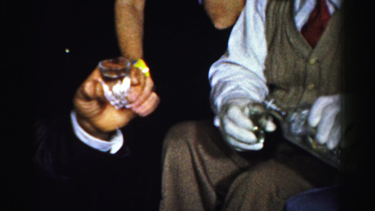 Adult hands hold a small glass while another seated man holds a bottle in a dim bar setting.