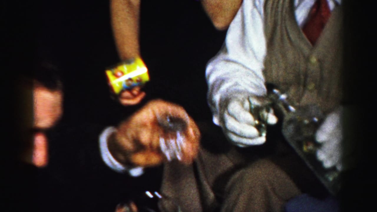 A hand extends a small glass while a seated man holds a bottle and another hand holds a yellow wrapper.