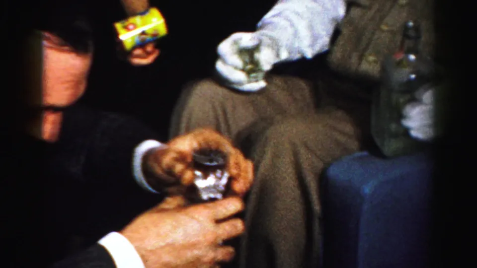 Close 1956 Albany home-movie view of hands with shot glasses, a seated man in vest and tie, and a bottle at the edge of frame.