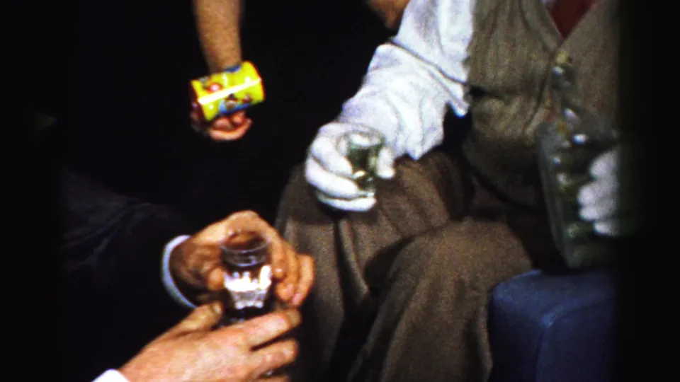 Albany 1956 frame showing adults around small shot glasses and a bottle, with vest, tie, trousers, and dim bar lighting visible.