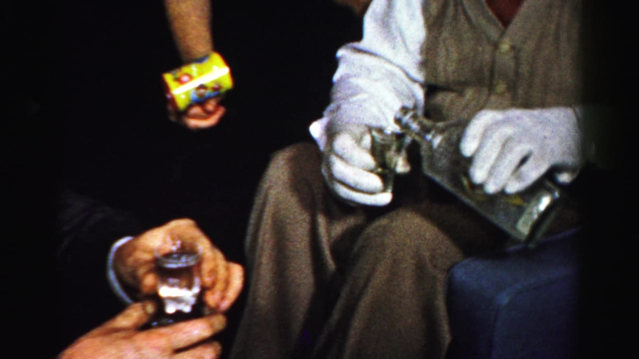 Albany-area 1950s home-movie frame showing a gloved man handling a bottle and glass beside a guest with a drink and snack package.
